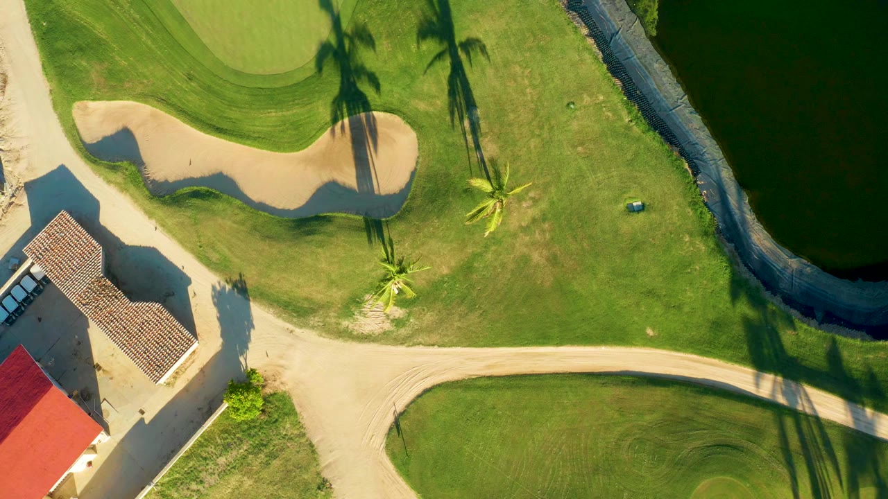 Second overhead frame of the San Jose Del Cabo golf course with the bunker and green at upper left, the water hazard along the right side, palm shadows crossing the grass, and no visible people.