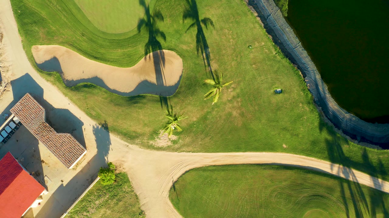 Mid-clip overhead view of the San Jose Del Cabo golf course with more of the red-roofed building visible at lower left, the bunker near the green, water to the right, long palm shadows, and no visible people.