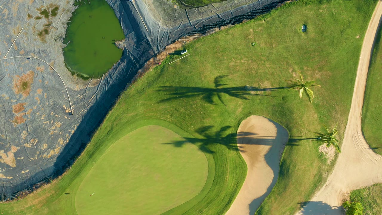Aerial frame in San Jose del Cabo showing the water hazard and black-lined ground at left, green grass across the center, the putting green low left, and the sand trap just right of it, with no visible people.