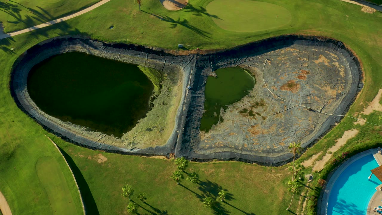 Overhead aerial frame in San Jose Del Cabo, Mexico showing two dark green lined ponds, bright grass, a curved path, palm shadows, and a partial blue swimming pool at the lower right.