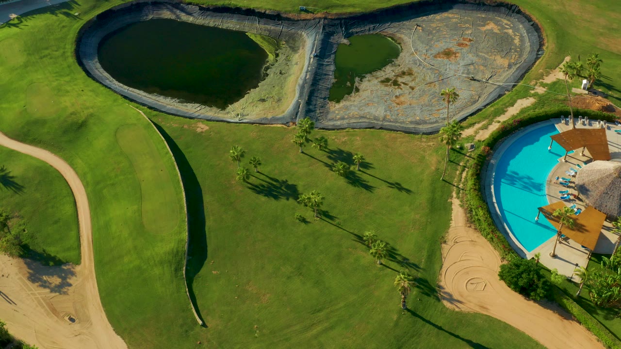 Overhead aerial frame in San Jose del Cabo showing a dark pond, a dry lined basin, green turf, curving paths, palm trees, and a blue pool at the right edge, with no visible people.