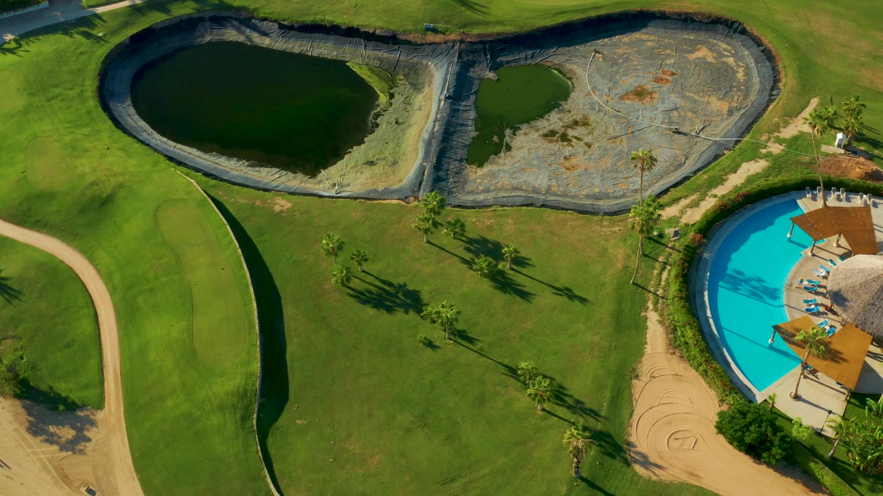 Overhead San Jose del Cabo frame where the dark water pond and pale dry pond basin fill the upper section, green turf fills the middle, and only part of the bright blue pool remains on the right, with no visible people.