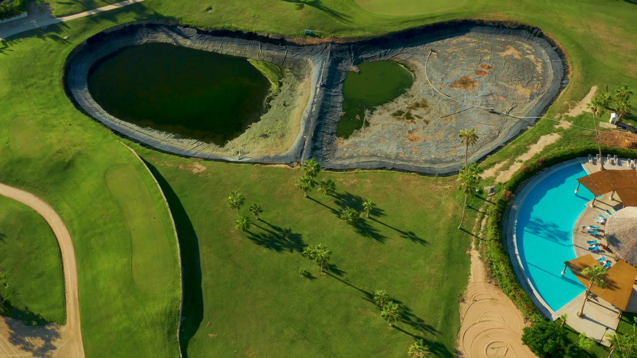 San Jose del Cabo drone frame with a dark pond at upper left, a dry basin at upper right, sweeping green grass below, path curves along the left and right, and a cropped blue pool at the lower right, with no visible people.