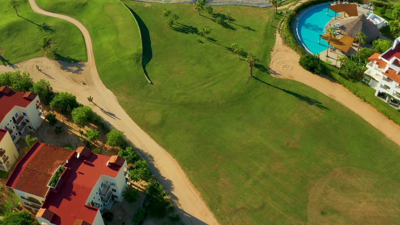 Aerial frame over a San Jose Del Cabo golf course showing green fairway, pale sand paths, red-roof buildings, a blue pool, trees, and no visible people.