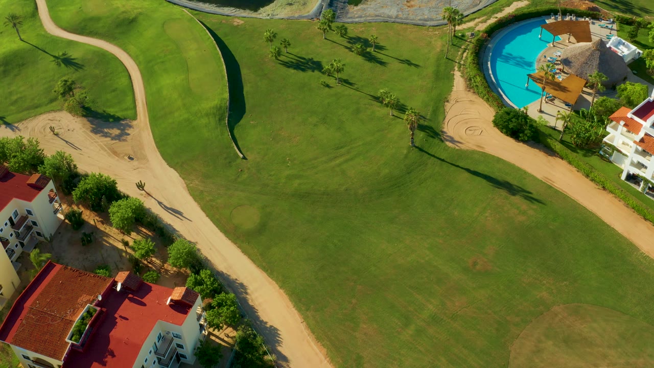 San Jose Del Cabo aerial frame showing golf greens, pale paths, a bright blue pool, roofed buildings, tree lines, and no visible people.