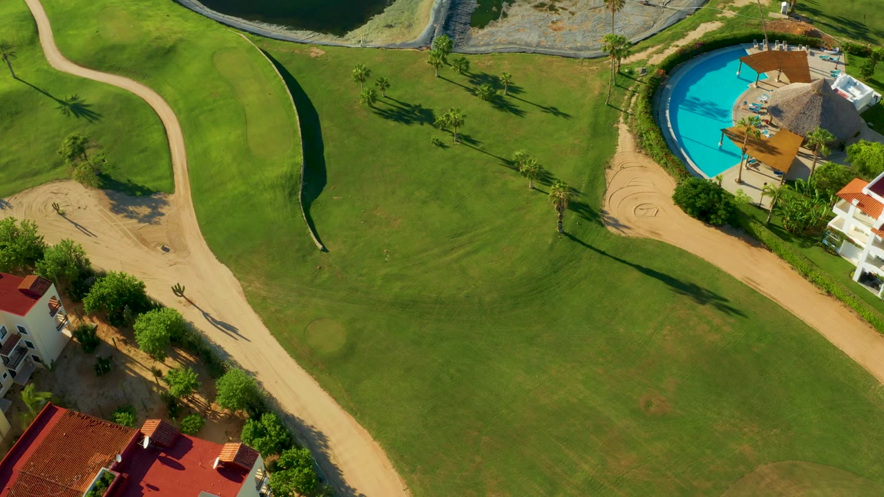 Aerial golf course frame with a pool, thatched roof, paths, and green fairway.