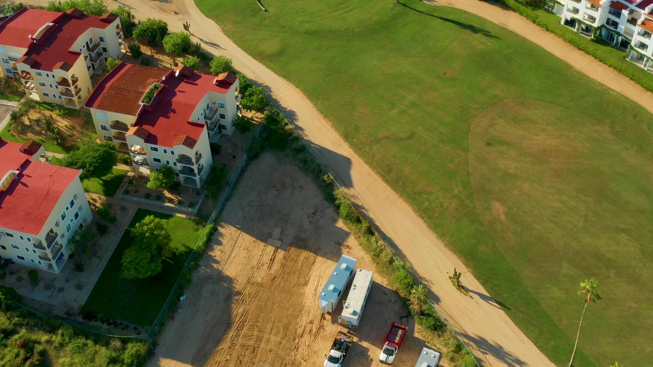 Aerial frame over San Jose Del Cabo showing red-roofed residential buildings, a diagonal dirt path, green grass, sandy ground, parked vehicles, trees, and no visible people.