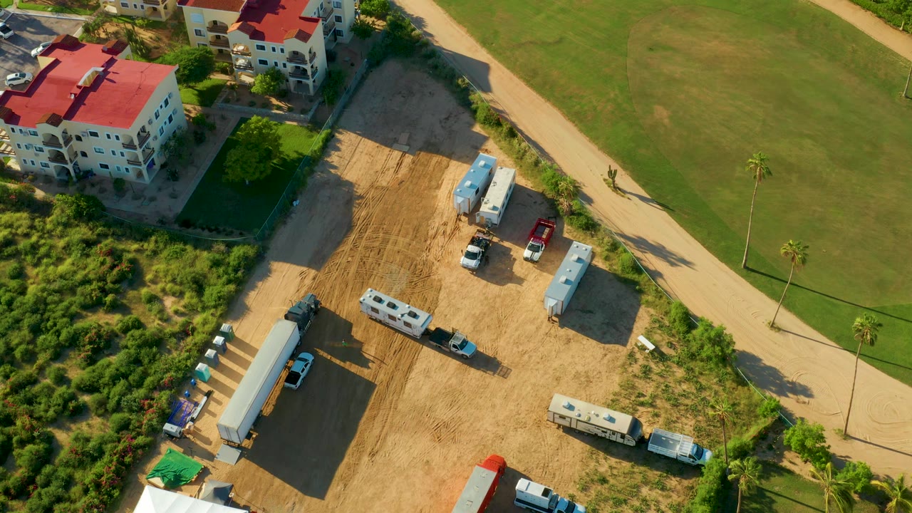 Aerial frame over San Jose Del Cabo showing a sandy construction lot, parked trailers and vehicles, red-roofed residential buildings, a pale road, green lawns, palms, and no visible people.