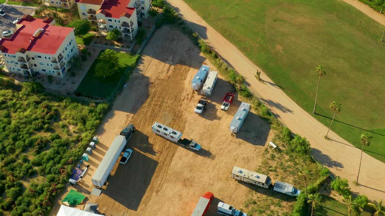 Drone view of construction vehicles and trailers near a road and lawns.