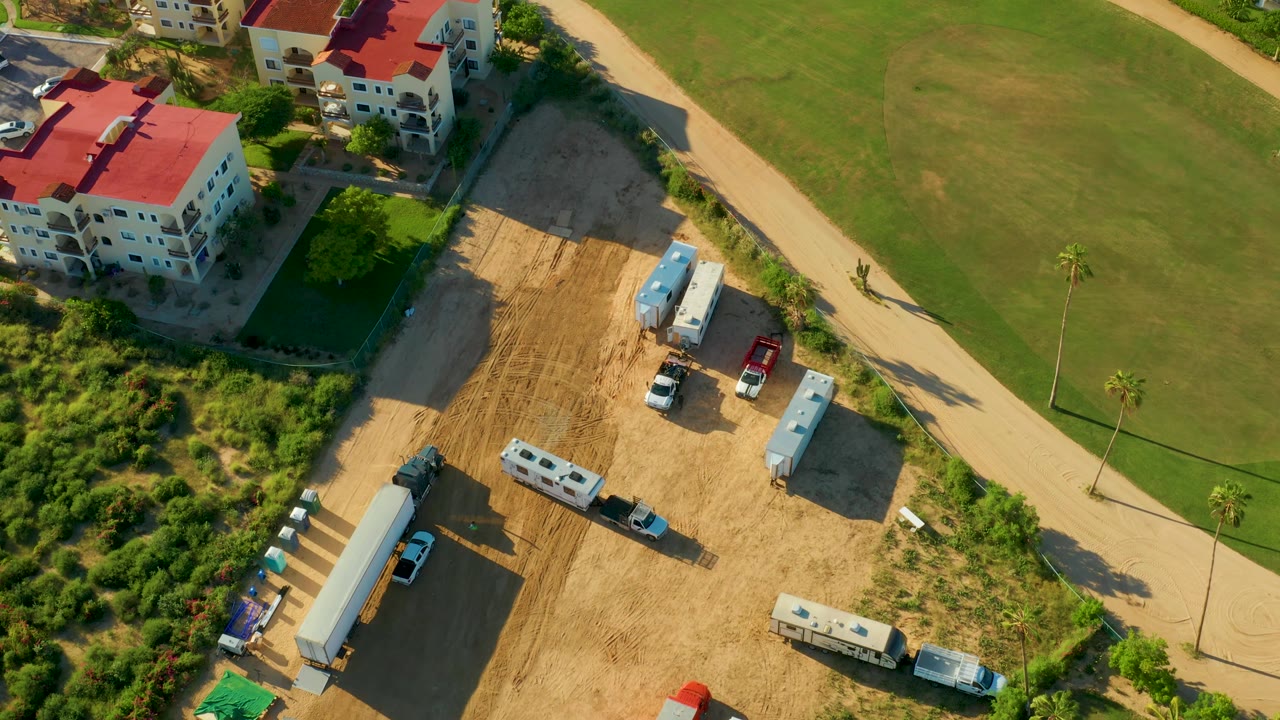 Aerial frame from San Jose Del Cabo with the red-roofed buildings higher in frame, the sandy lot below them, parked work vehicles and trailers, a curved pale road, green grass, and no visible people.