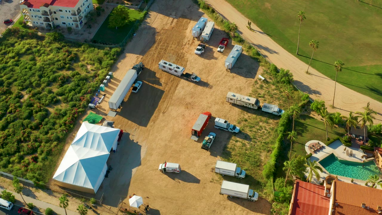 Aerial frame from San Jose Del Cabo showing a white tent, trailers, trucks, a red-roofed building edge, green grass, pathways, palm trees, and a pool area with no visible people.