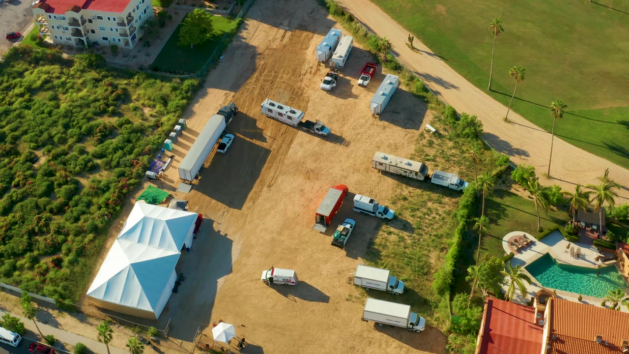 San Jose Del Cabo aerial still with the dirt staging lot centered, a white tent at lower left, service vehicles and trailers across the lot, and landscaped grass and pool at the right with no visible people.