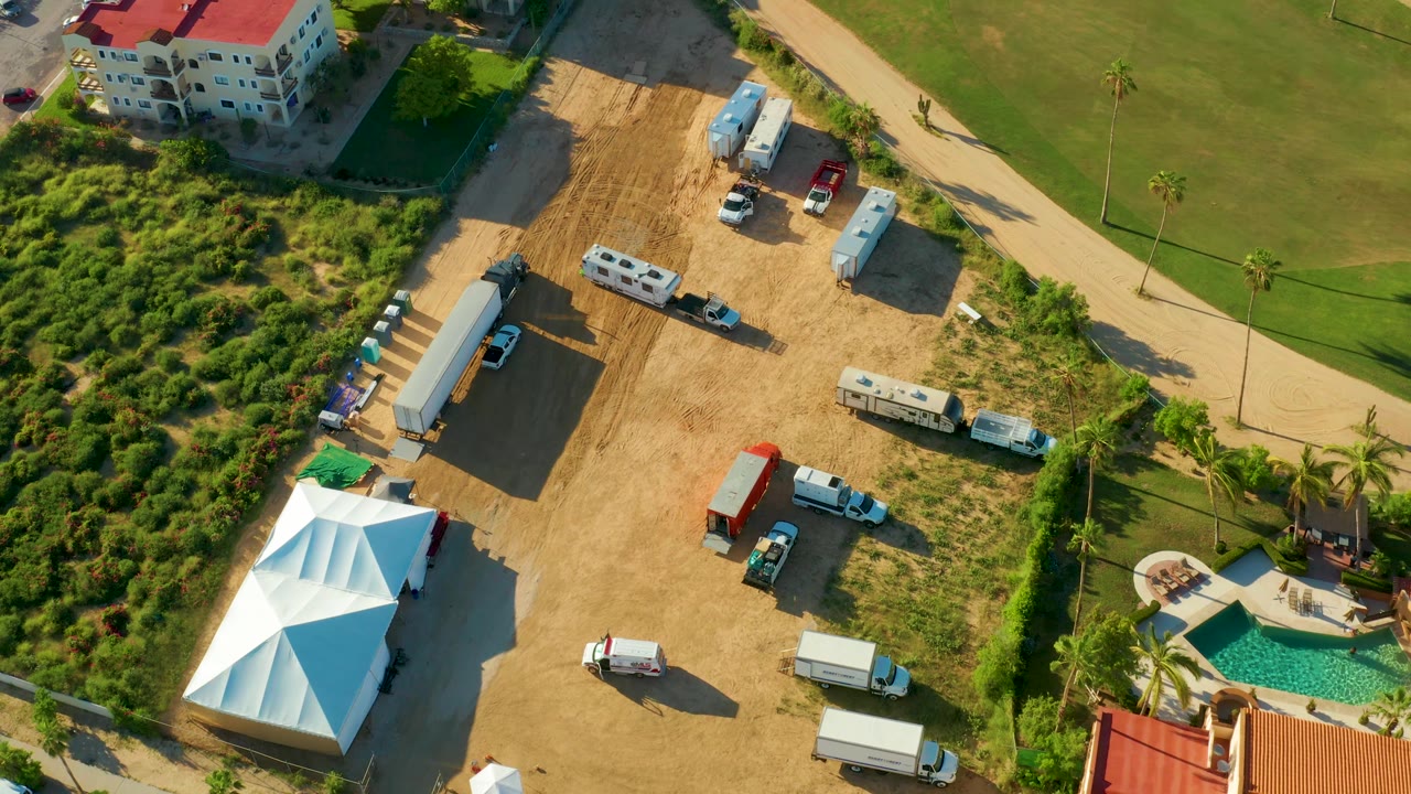 Overhead San Jose Del Cabo frame showing the white tent partly at lower left, multiple trucks and trailers on the dirt lot, a red-roofed building edge, a lawn, a curved pathway, palms, and no visible people.