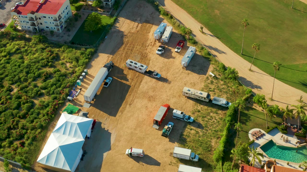 High aerial view of a temporary lot with tents, trucks, grass, palms, and nearby buildings.