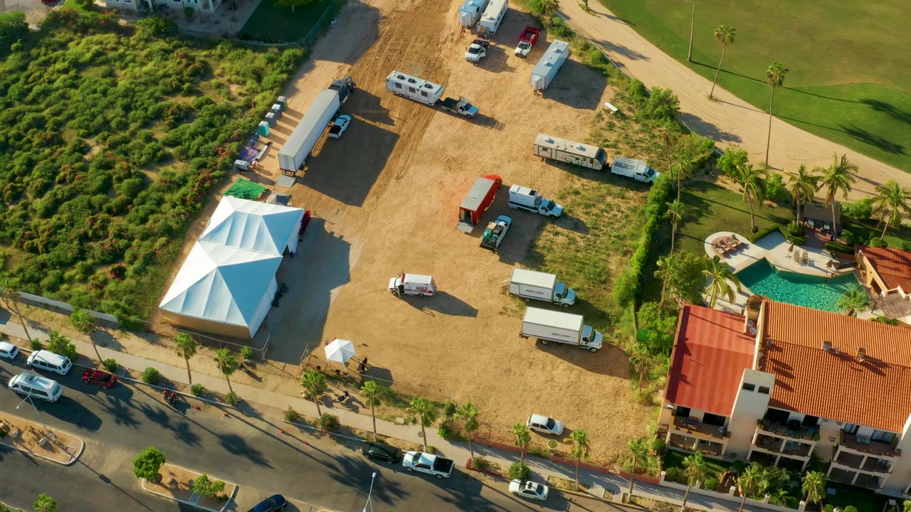 Drone view of a dirt lot with parked vehicles next to greenery, road, buildings, and pool.