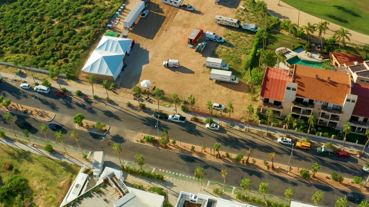 Aerial frame from San Jose del Cabo showing a palm-lined multi-lane road, cars, a dirt lot with parked trucks, a white tent, red-roofed buildings, greenery, and no visible people.