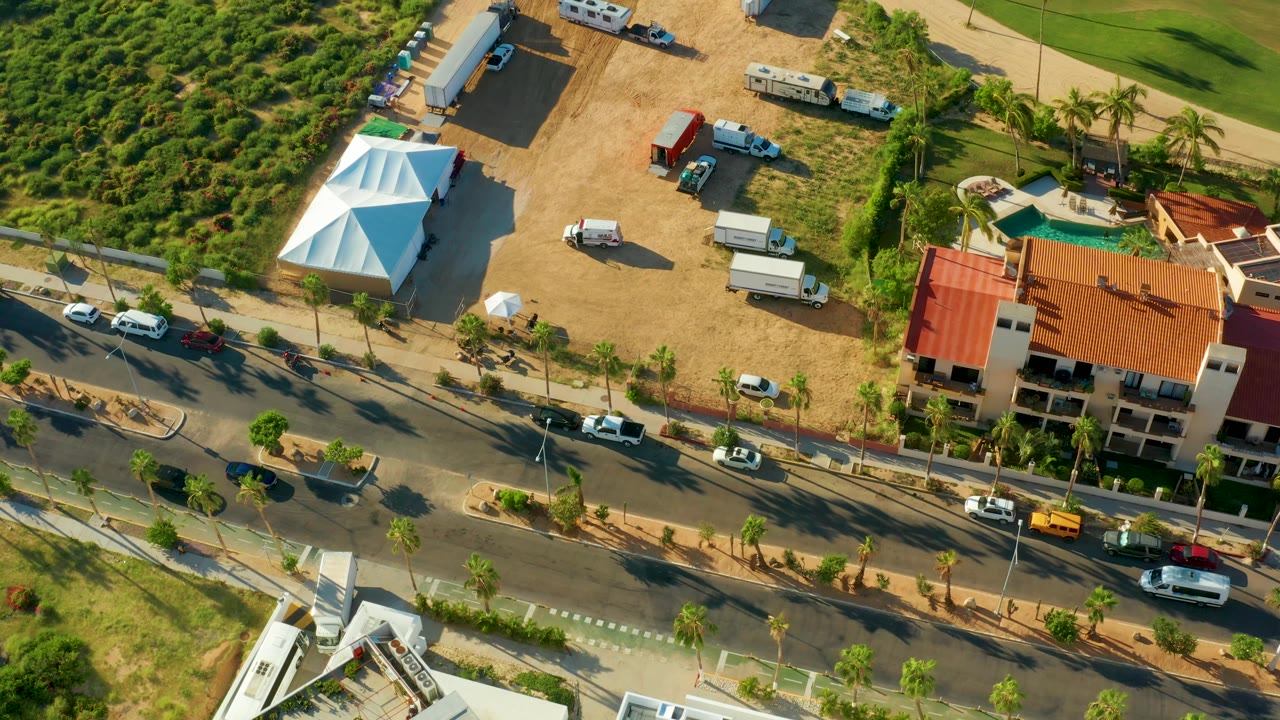 San Jose del Cabo aerial frame showing the divided road with cars and vans, planted medians, palm trees, a white tent, dirt parking area, red-tile roof building, and no visible people.