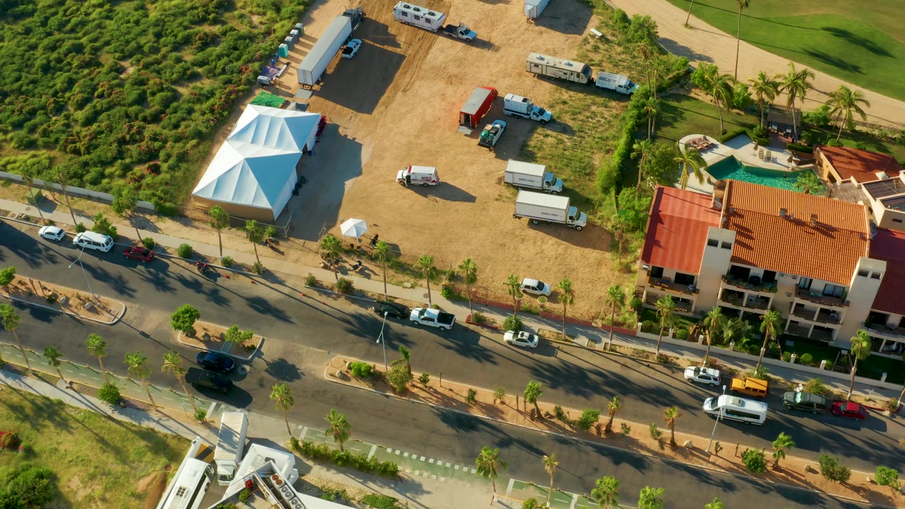 Aerial shot of a dirt lot with a white tent above a palm-lined traffic road.