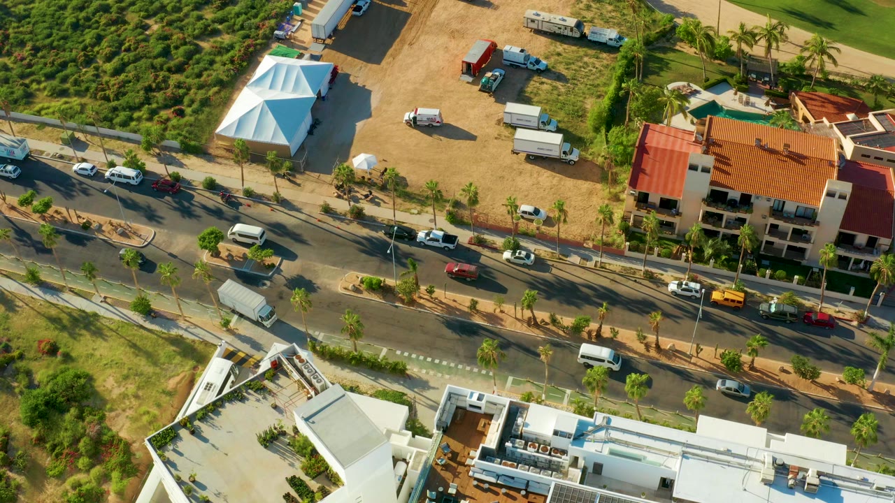 San Jose Del Cabo aerial view with cars and vans spread along the palm-lined road, the white tent at upper left, the red-roofed building at right, and no visible people.