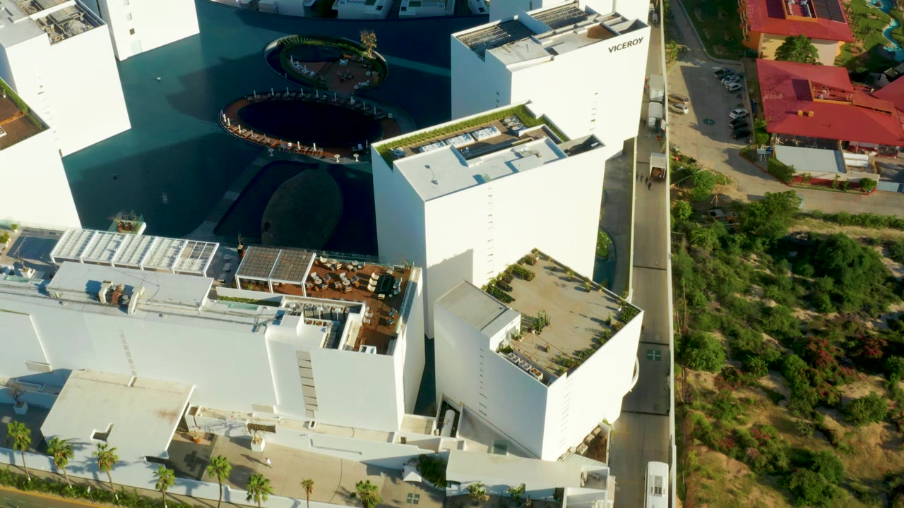 Aerial frame over a modern residential complex in San Jose Del Cabo, with white buildings, flat roofs, a dark blue water feature, roads, greenery, and no visible people.