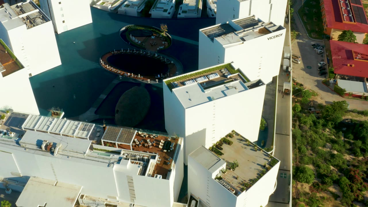Aerial frame over the San Jose Del Cabo complex with white residential blocks, roof terraces, a dark blue central pool or lagoon feature, roadside greenery, and no visible people.