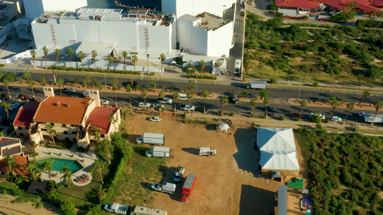 Aerial frame over San Jose Del Cabo showing a palm-lined road between a large white building, a red-roof house, a pool, dry lots, and no visible people.