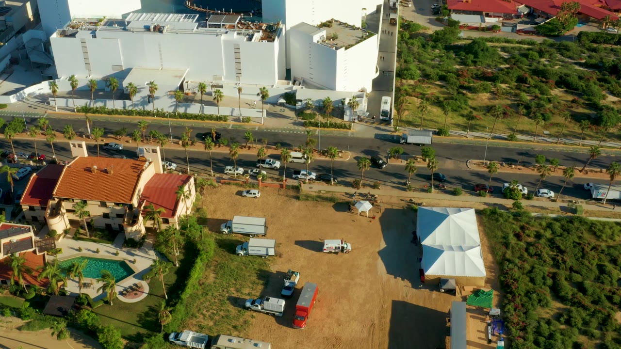 Aerial San Jose Del Cabo view with more of the white building and hillside visible above the road, while the red-roof home, pool, vehicles, and no visible people remain in frame.