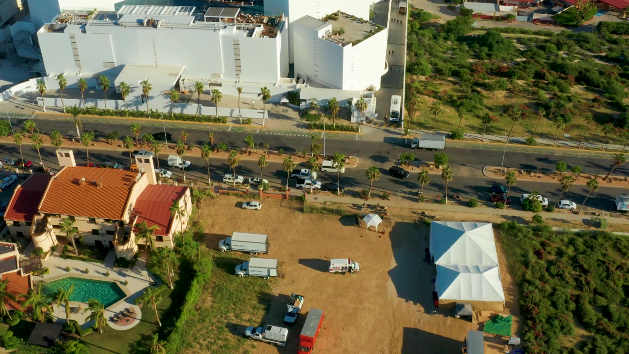 High oblique aerial frame in San Jose Del Cabo showing the road with several vehicles, the white building complex, the red-roof residence, a pool corner, dry ground, and no visible people.