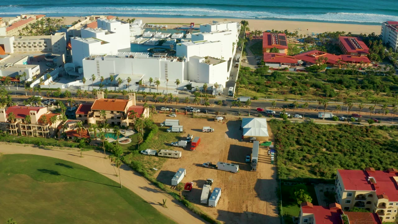 Aerial frame over San Jose del Cabo showing the ocean and beach across the top, large white buildings, red-roofed structures, a road with cars, green lots, and no visible people.
