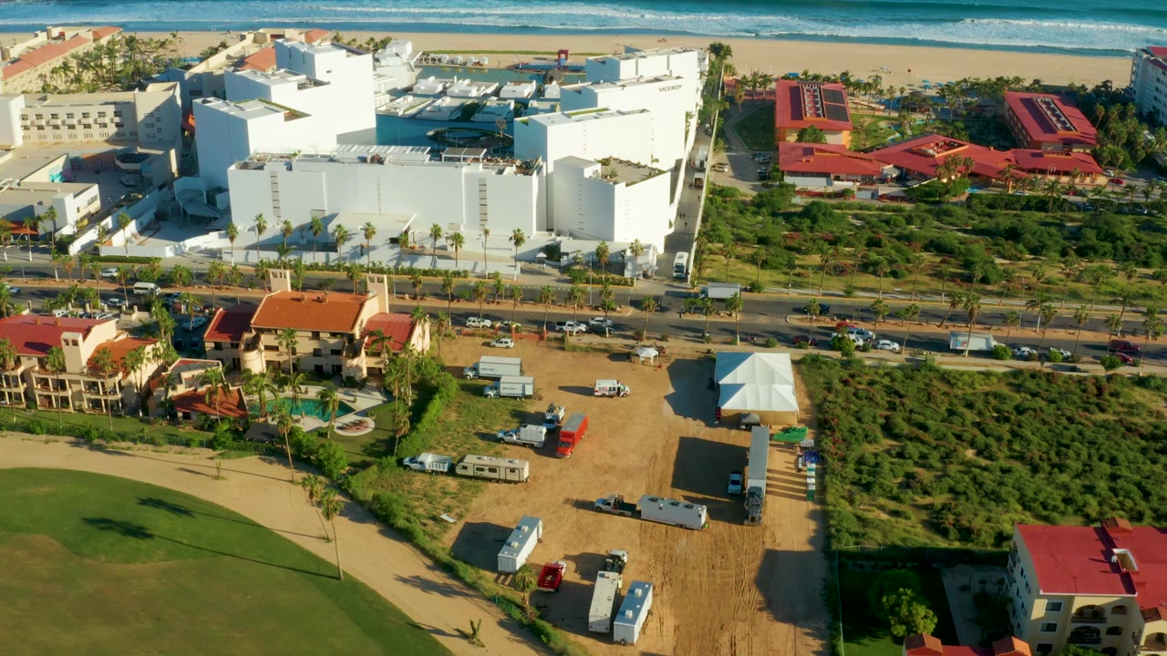 San Jose del Cabo frame with a bright white building complex near center, red-roofed buildings on the right, a road with cars below, a green undeveloped parcel, beach and ocean at the top, and no visible people.