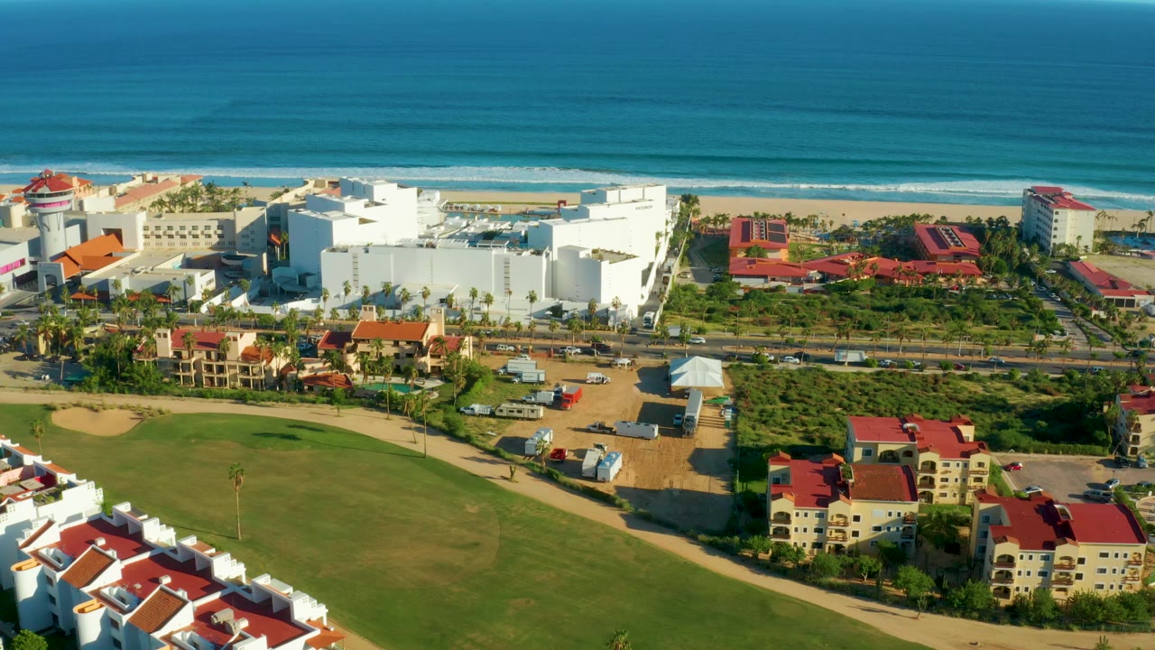 Opening aerial frame over San Jose Del Cabo with blue ocean and sandy beach behind white coastal buildings, red roofs, road, green lawn, and no visible people.
