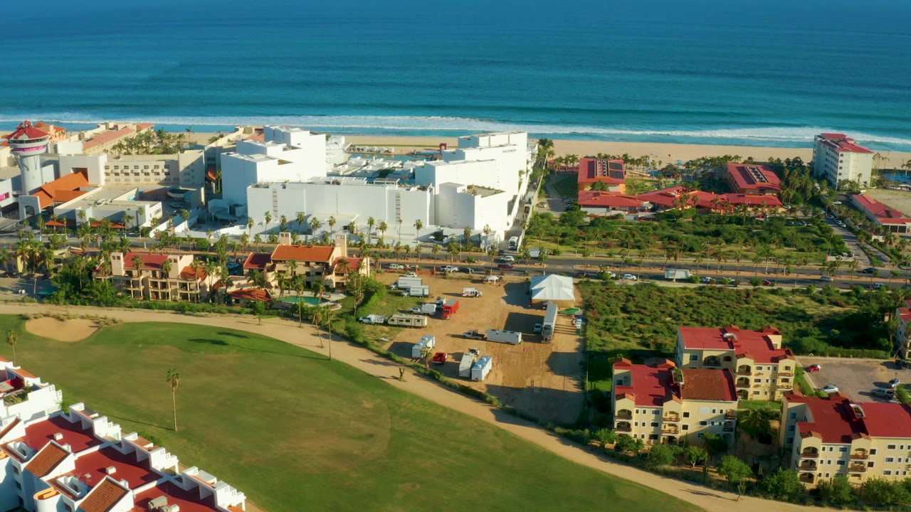 Two seconds into the San Jose Del Cabo pan, the white coastal buildings and red-roofed properties sit between the beach and a broad green lawn, with no visible people.