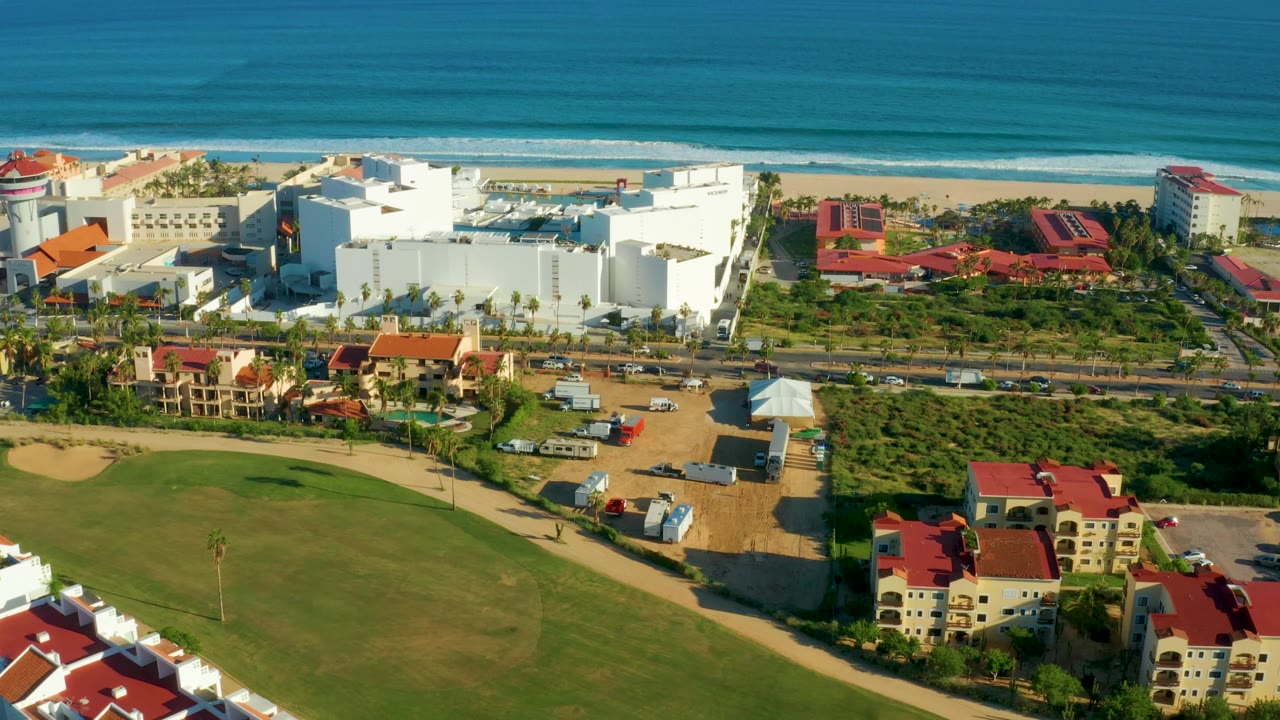 Drone view of beach, ocean, white coastal buildings, red roofs, road, palms, and green grounds.