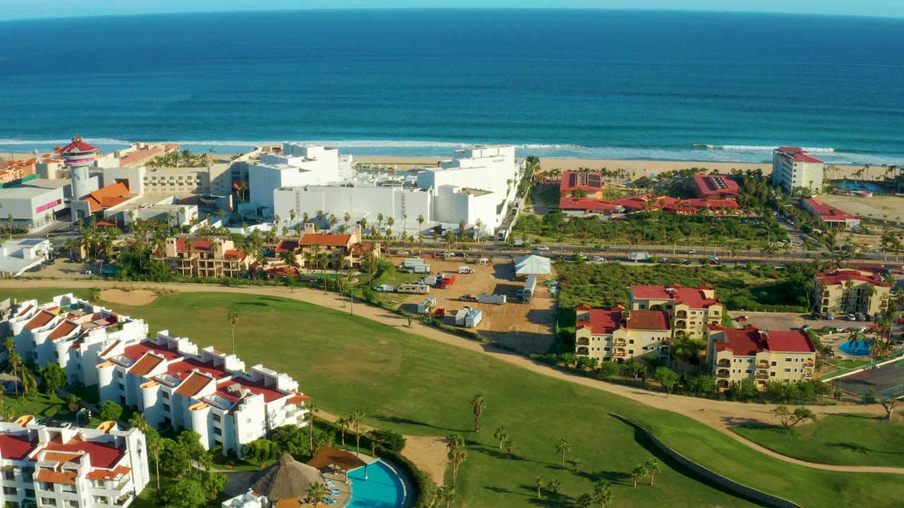 A slightly later San Jose del Cabo aerial view keeps the ocean horizon high while white buildings, red roofs, roads, palms, and green resort lawns fill the lower frame with no visible people.