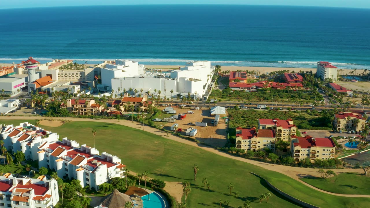 Mid-clip aerial frame in San Jose del Cabo with green lawns in the foreground, white resort buildings and red roofs in the middle distance, beach and ocean behind, and no visible people.