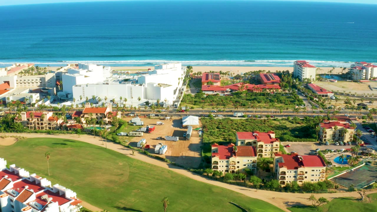 Opening aerial view in San Jose Del Cabo with blue ocean at the horizon, sandy beach, white resort buildings, red roofs, green lawns, and no visible people.