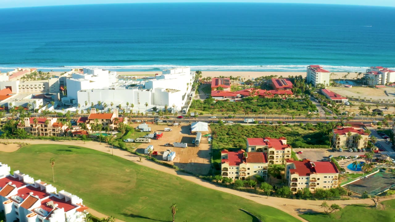 Aerial view over San Jose Del Cabo resort grounds with red roofs and white buildings in the lower half, beach and ocean stretching across the upper half, and no visible people.