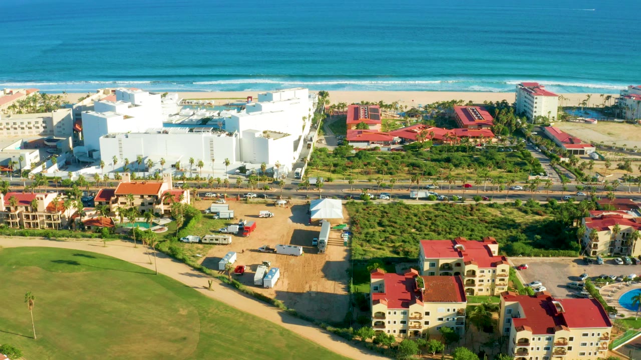 Aerial view over San Jose Del Cabo with blue ocean and surf at the top, white resort buildings and red roofs near the beach, a road with cars, green golf-course grass in the foreground, and no visible people.