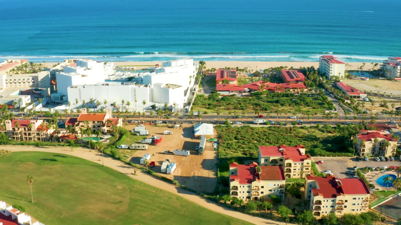 Aerial beach scene with ocean, white hotel-like buildings, red roofs, cars, and golf grass