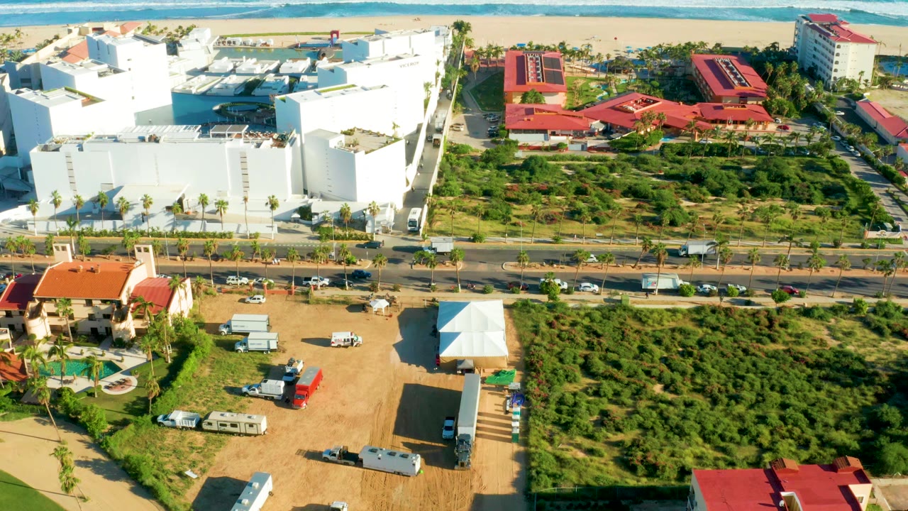 Opening aerial frame over San Jose del Cabo with white beachfront buildings at left, red-roofed structures near the beach, a road with cars, green lots in the foreground, ocean surf beyond, and no visible people.