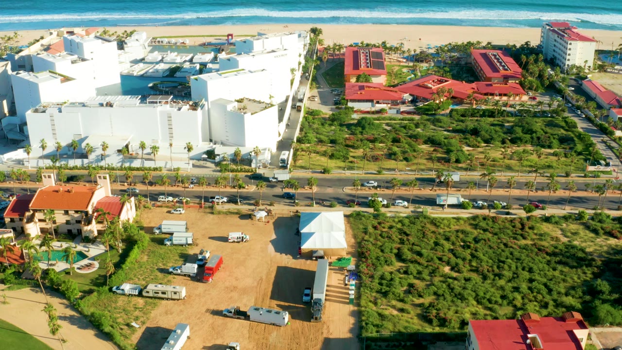 San Jose del Cabo drone frame looking across open green parcels and a roadside strip toward white oceanfront buildings, red roofs, sandy beach, blue surf, parked vehicles, and no visible people.
