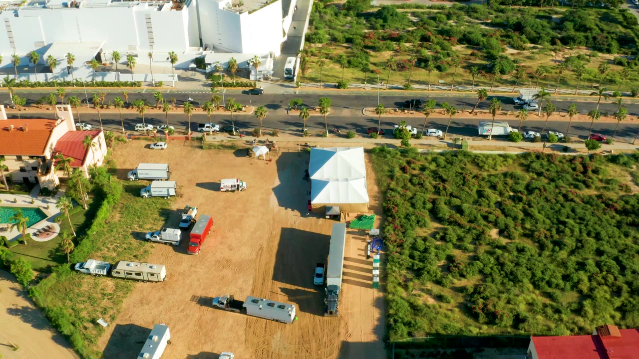 Aerial frame from San Jose Del Cabo showing a divided road with cars, a dirt parking lot with vehicles and a white tent, red-roofed homes, green shrubs, and no visible people.