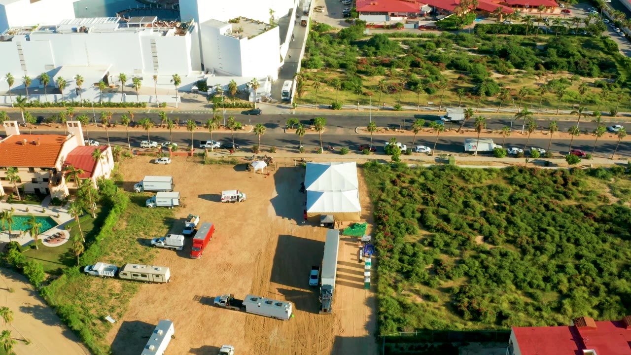 Roadside aerial with cars, a large white building, a green lot, and residential roofs.