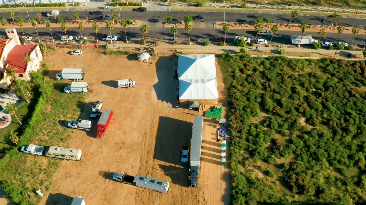 Overhead frame in San Jose Del Cabo showing a dirt lot with RVs, trucks, a white tent, a red-roofed building edge, a road with cars, dense greenery, and no visible people.