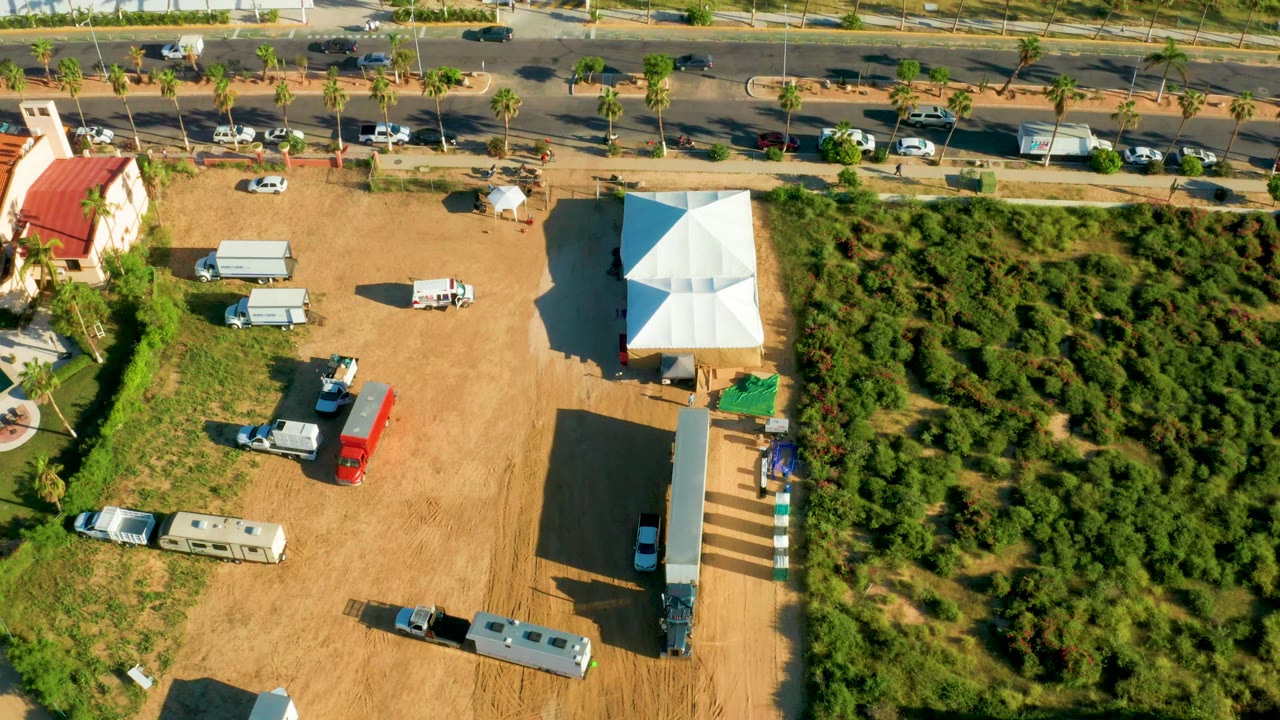 Aerial frame in San Jose Del Cabo with RVs and utility vehicles parked on a dirt lot, a white tent near the center-right, traffic along the road, greenery to the right, and no visible people.