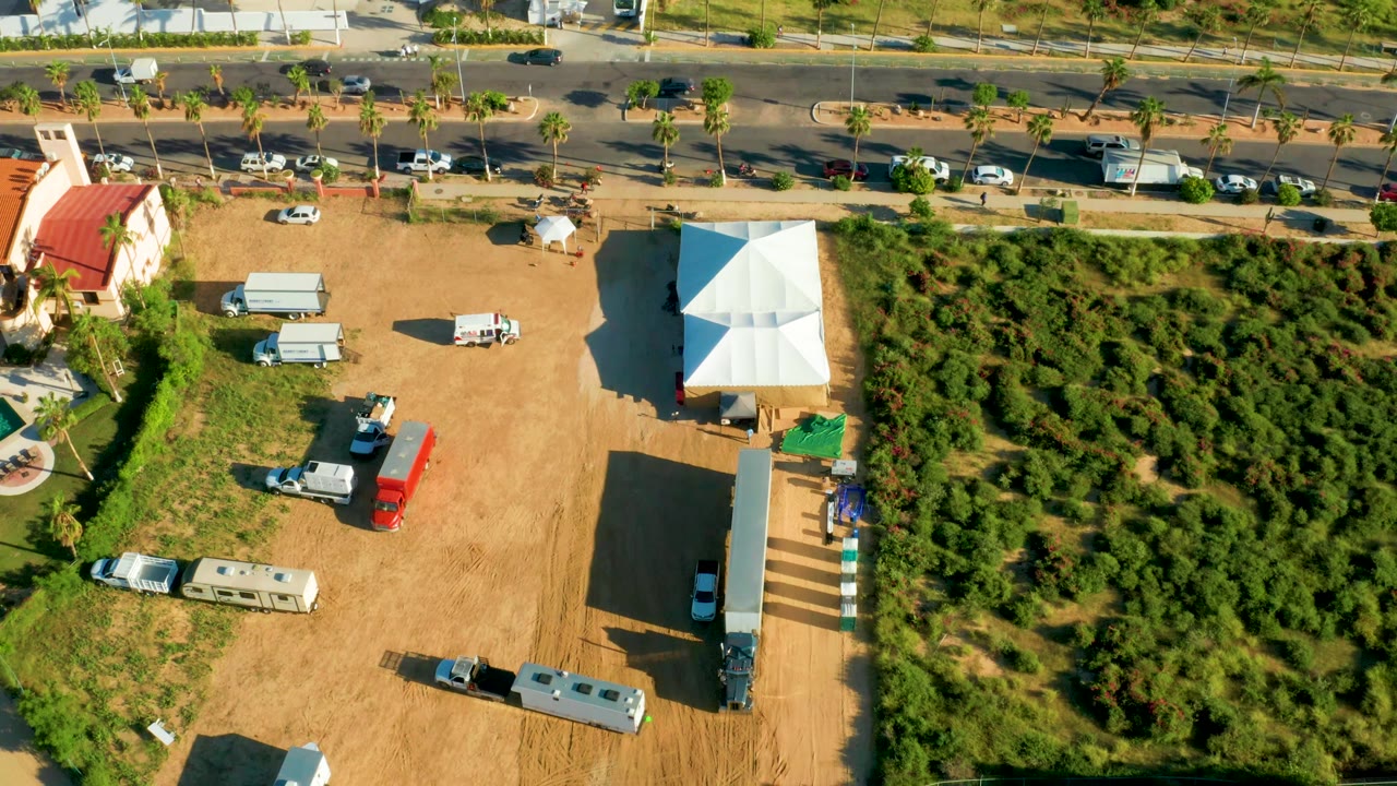 Dirt lot with white tent, parked vehicles, greenery, and road traffic