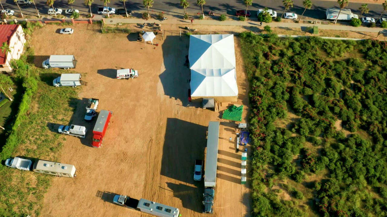 Overhead view in San Jose Del Cabo showing a paved road, dirt lot, white tents, RVs, parked vehicles, palms, and dense green brush, with no visible people.