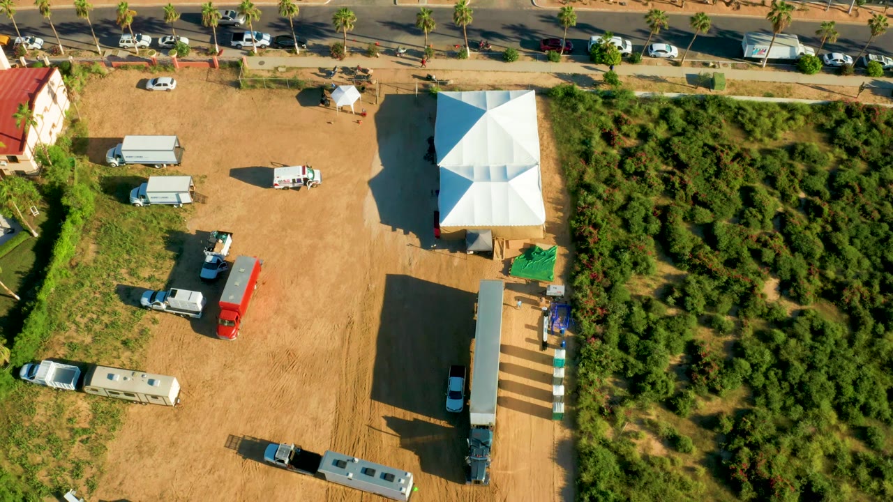 Roadside dirt lot with white tents, RVs, vans, and palm-lined road