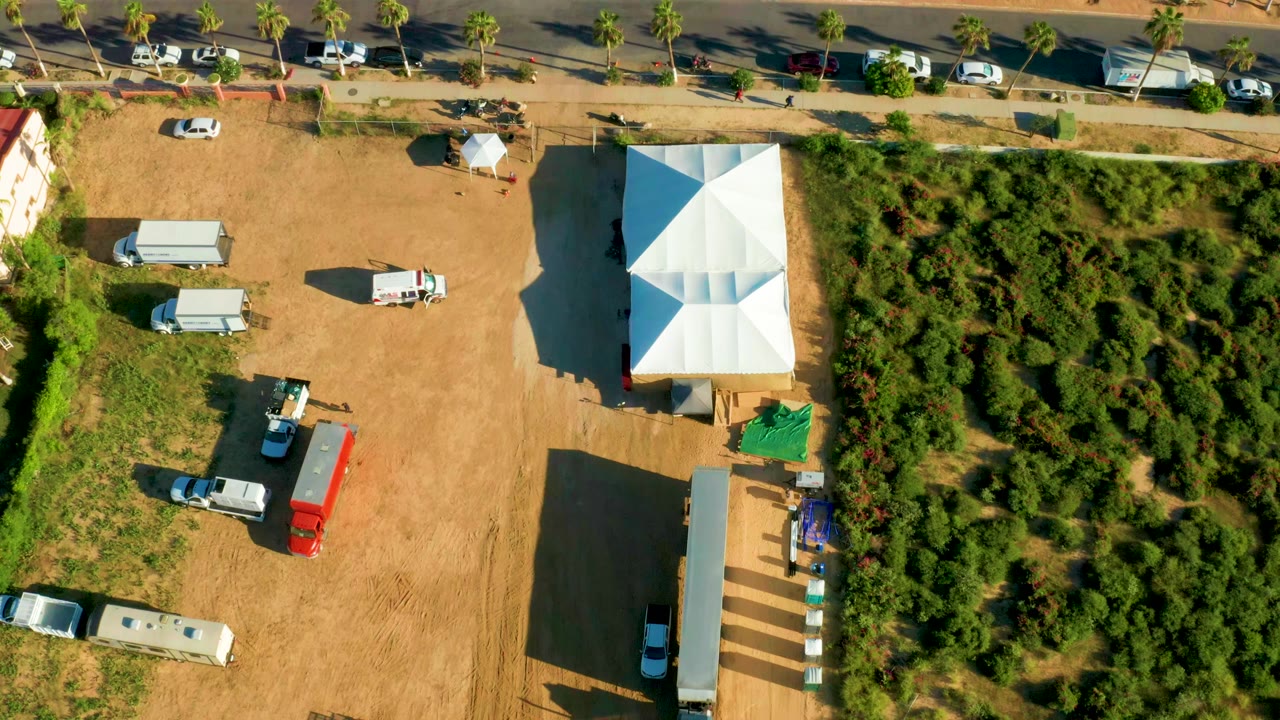 Aerial view of vehicles spaced across a dirt lot beside a white tent and dense bushes.