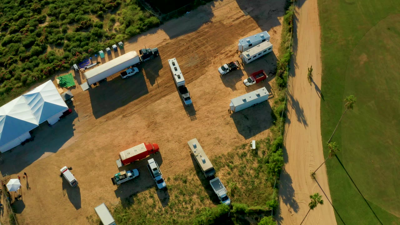 Aerial view in San Jose Del Cabo showing white tents, RVs, parked vehicles, dirt ground, a grassy edge, and a road at the start of the clip, with no clearly visible people.